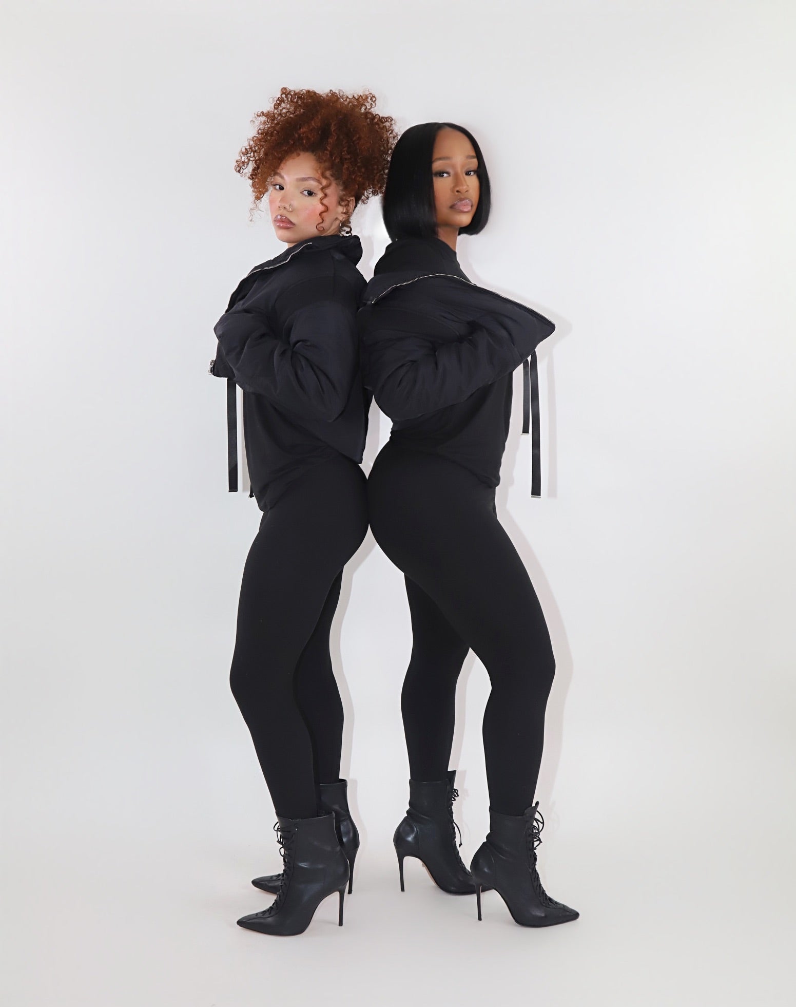 Woman with curly red hair wearing a black cropped puffer jacket over a matching black fitted outfit, posing with hands on the jacket collar against a plain white background. Trendy and cozy outerwear look