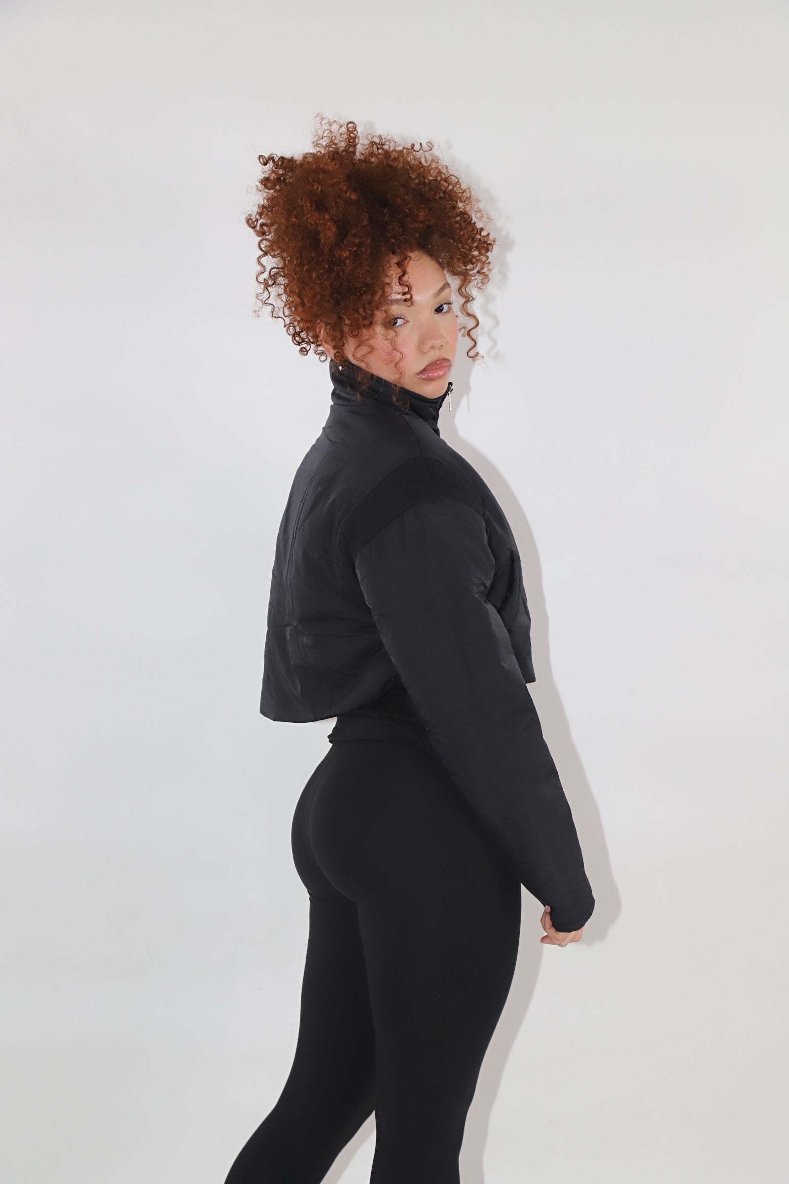 Woman with curly red hair wearing a black cropped puffer jacket over a matching black fitted outfit, posing with hands on the jacket collar against a plain white background. Trendy and cozy outerwear look