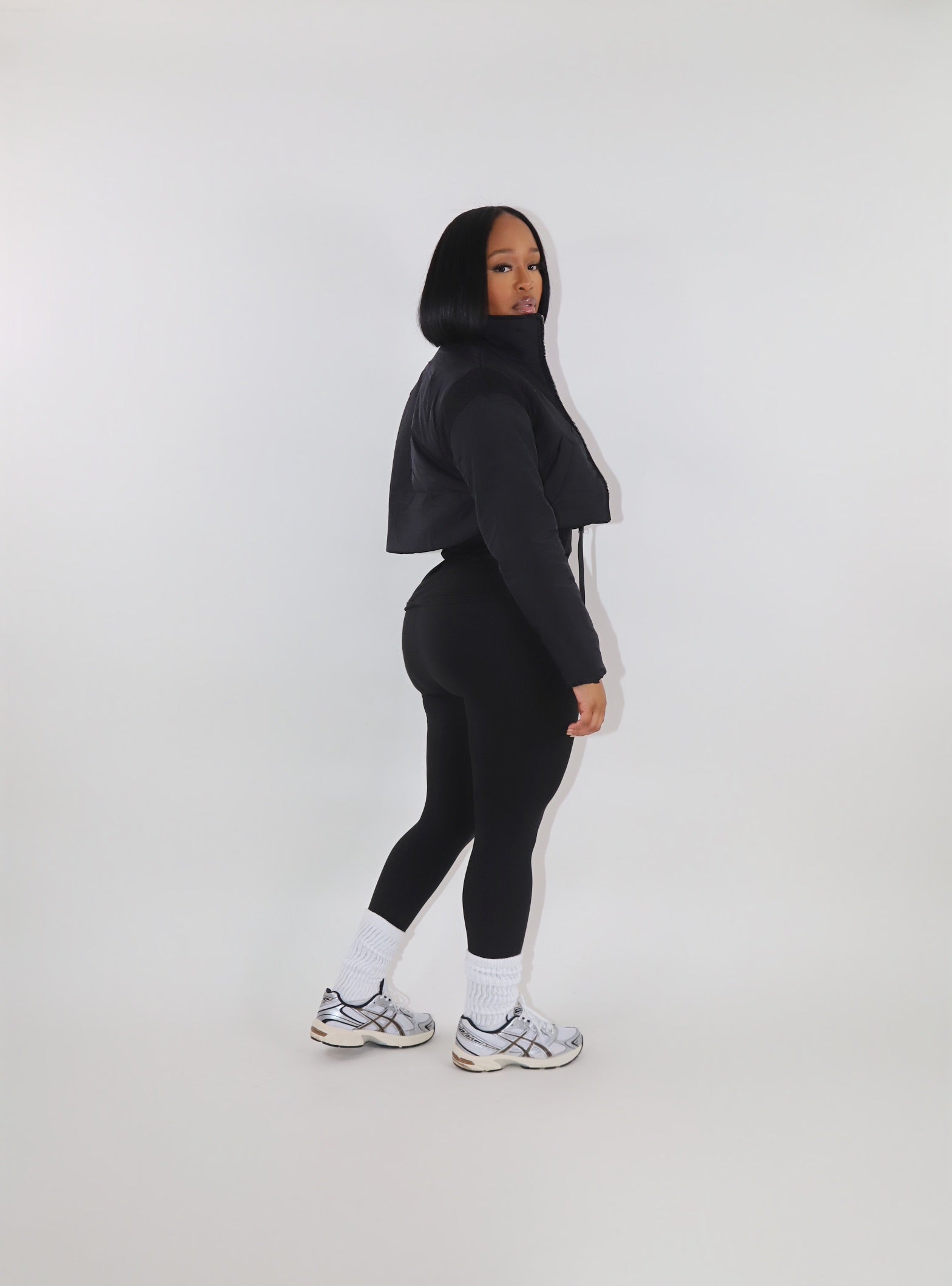 Woman with curly red hair wearing a black cropped puffer jacket over a matching black fitted outfit, posing with hands on the jacket collar against a plain white background. Trendy and cozy outerwear look-get the look at Ehrow Studios