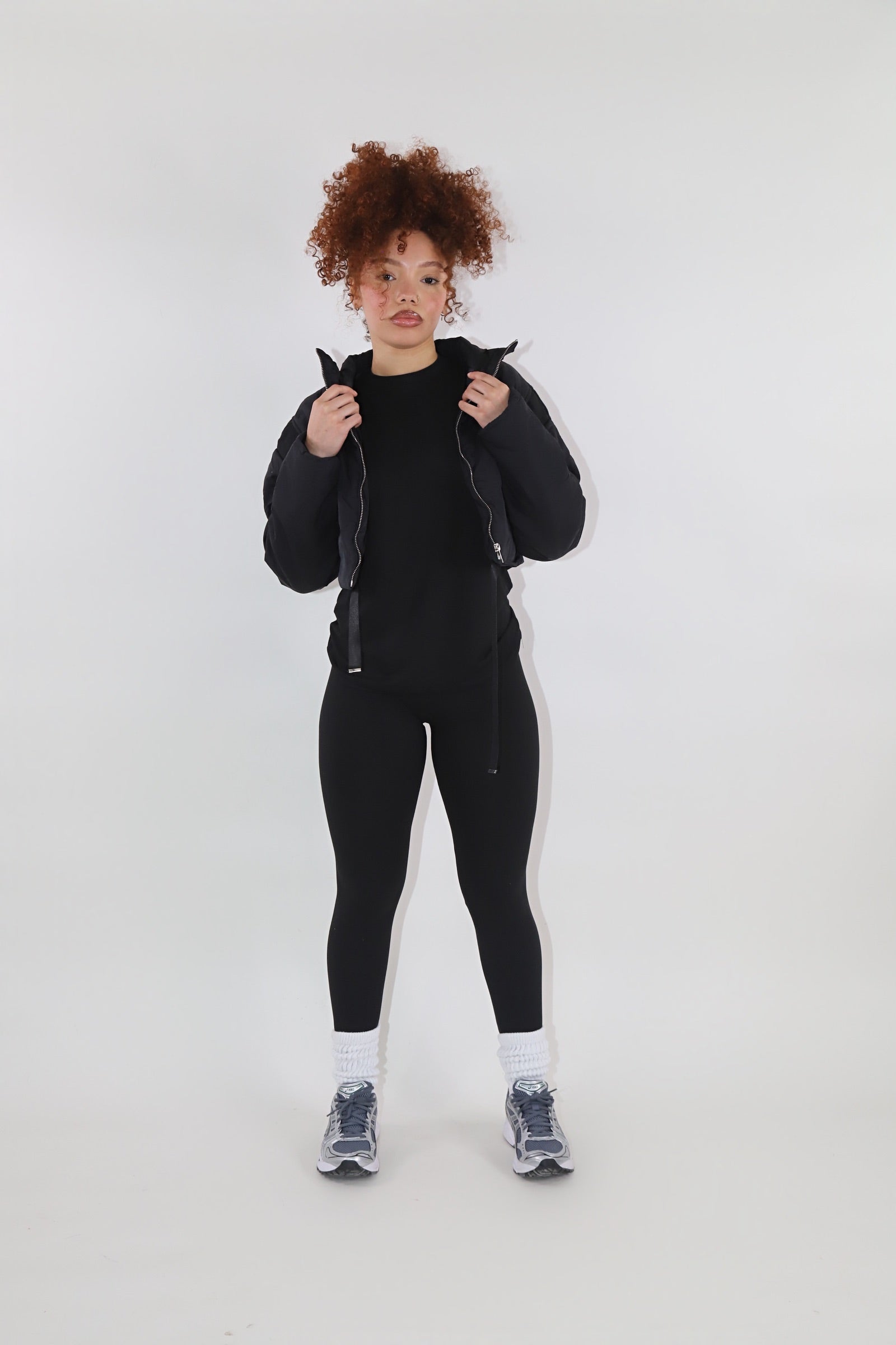 Woman with curly red hair wearing a brown cropped puffer jacket over a matching brown fitted outfit, posing with hands on the jacket collar against a plain white background. Trendy and cozy outerwear loo