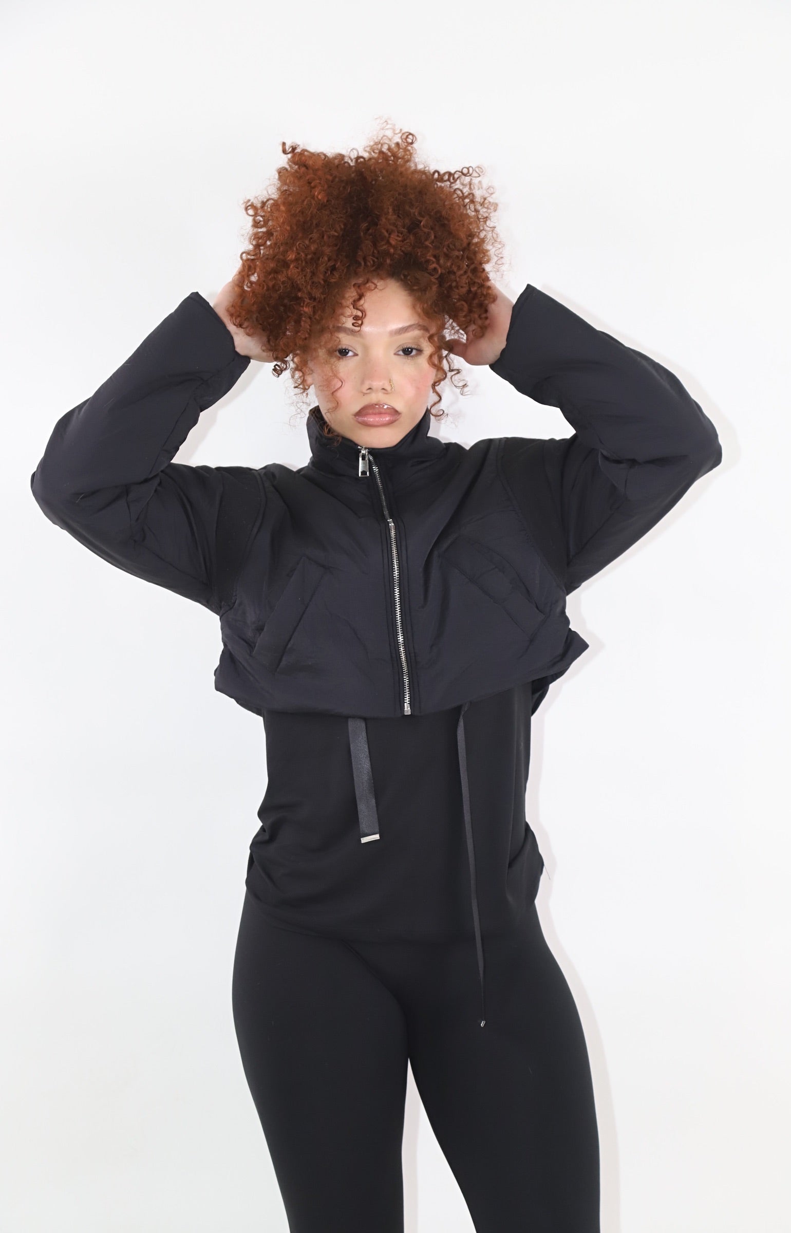 Woman with curly red hair wearing a black cropped puffer jacket over a matching black fitted outfit, posing with hands on the jacket collar against a plain white background. Trendy and cozy outerwear look-get the look at Ehrow Studios