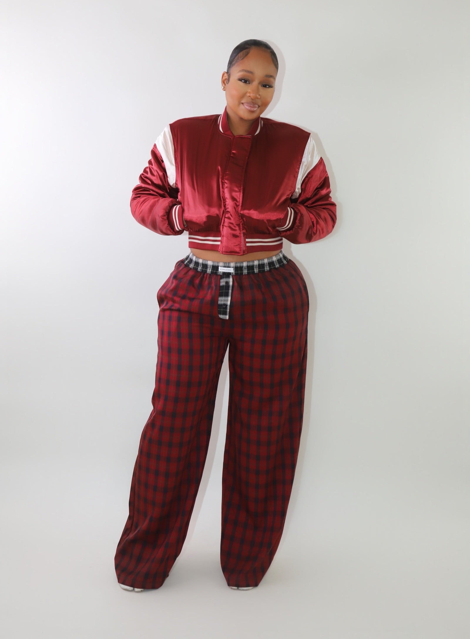 A woman wearing a cropped red satin bomber jacket with white sleeve accents and striped ribbed cuffs. The jacket has a varsity-inspired design with a sleek, shiny finish, giving it a sporty yet stylish look.