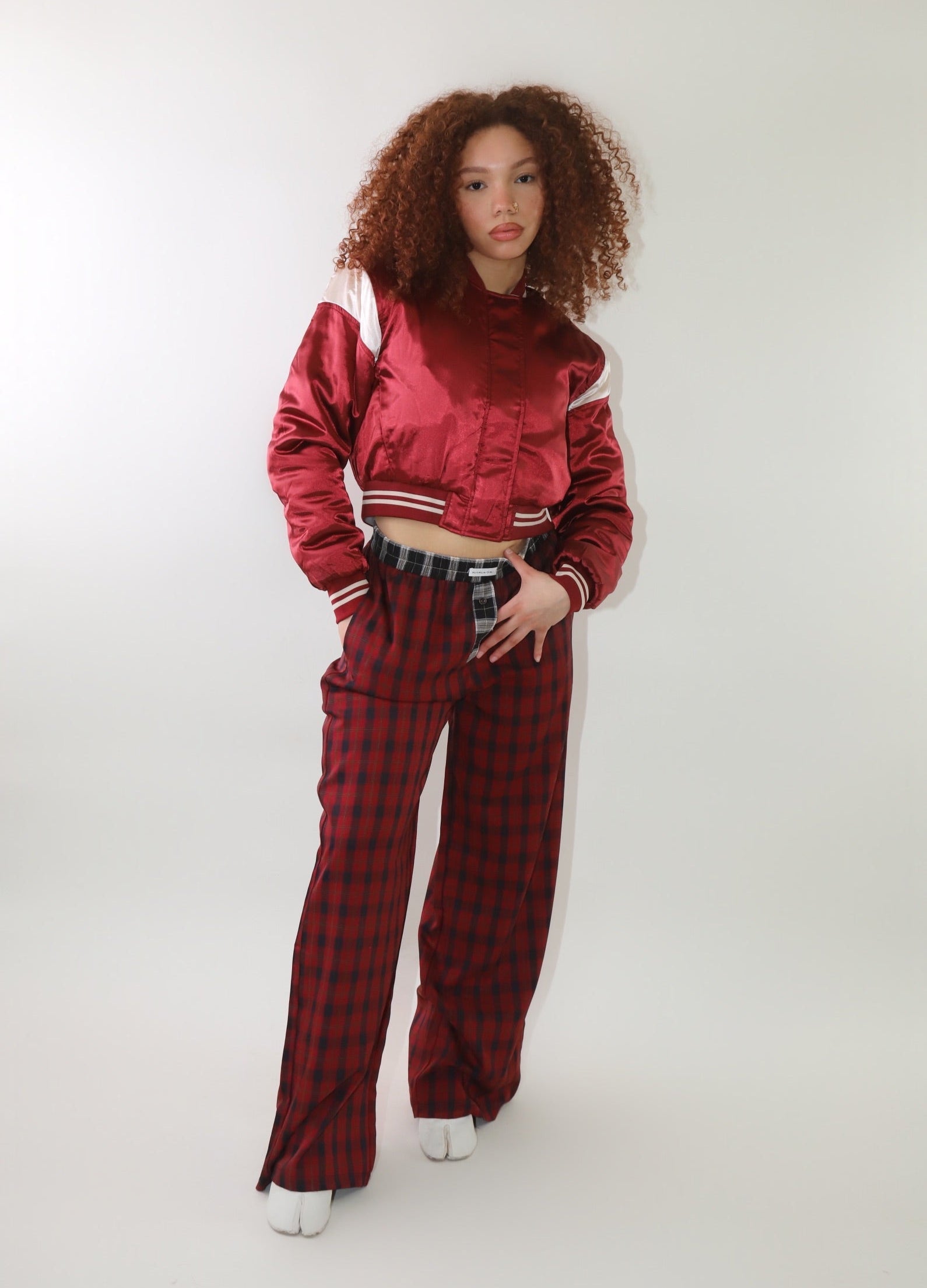 A woman wearing a cropped burgundy satin bomber jacket with white sleeve accents and striped ribbed cuffs. The jacket has a varsity-inspired design with a sleek, shiny finish, giving it a sporty yet stylish look.