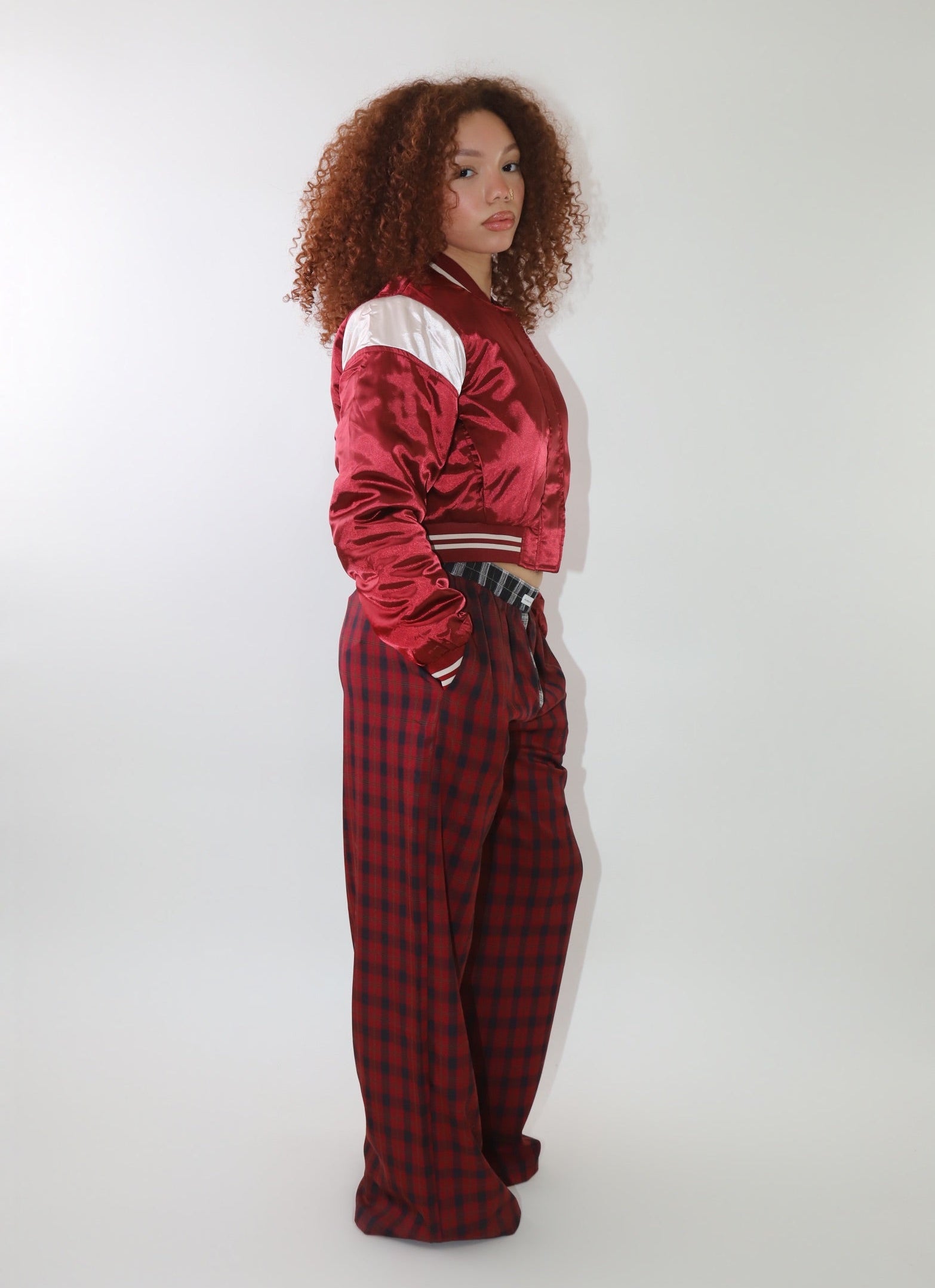 A woman wearing a cropped burgundy satin bomber jacket with white sleeve accents and striped ribbed cuffs. The jacket has a varsity-inspired design with a sleek, shiny finish, giving it a sporty yet stylish look.
