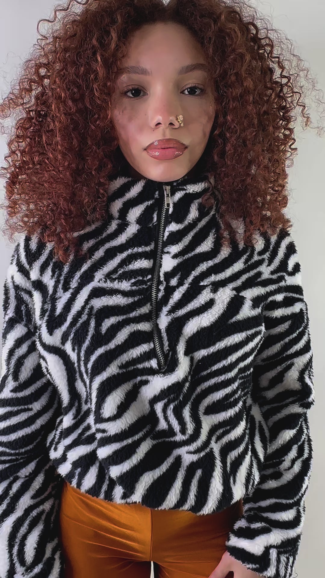 A woman with voluminous curly red hair wears a black-and-white zebra print Sherpa pullover with a high collar and half-zip closure. She pairs it with burnt-orange leggings, standing against a white background showing how the zebra sherpa fleece fits