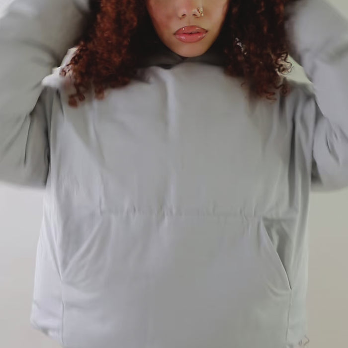 A woman with curly red hair wears a light gray oversized puffer jacket hoodie with a relaxed fit and kangaroo pockets. She pairs it with burnt-orange leggings, standing against a plain white background. The hoodie has a minimal design with a soft, padded texture, ideal for warmth and style