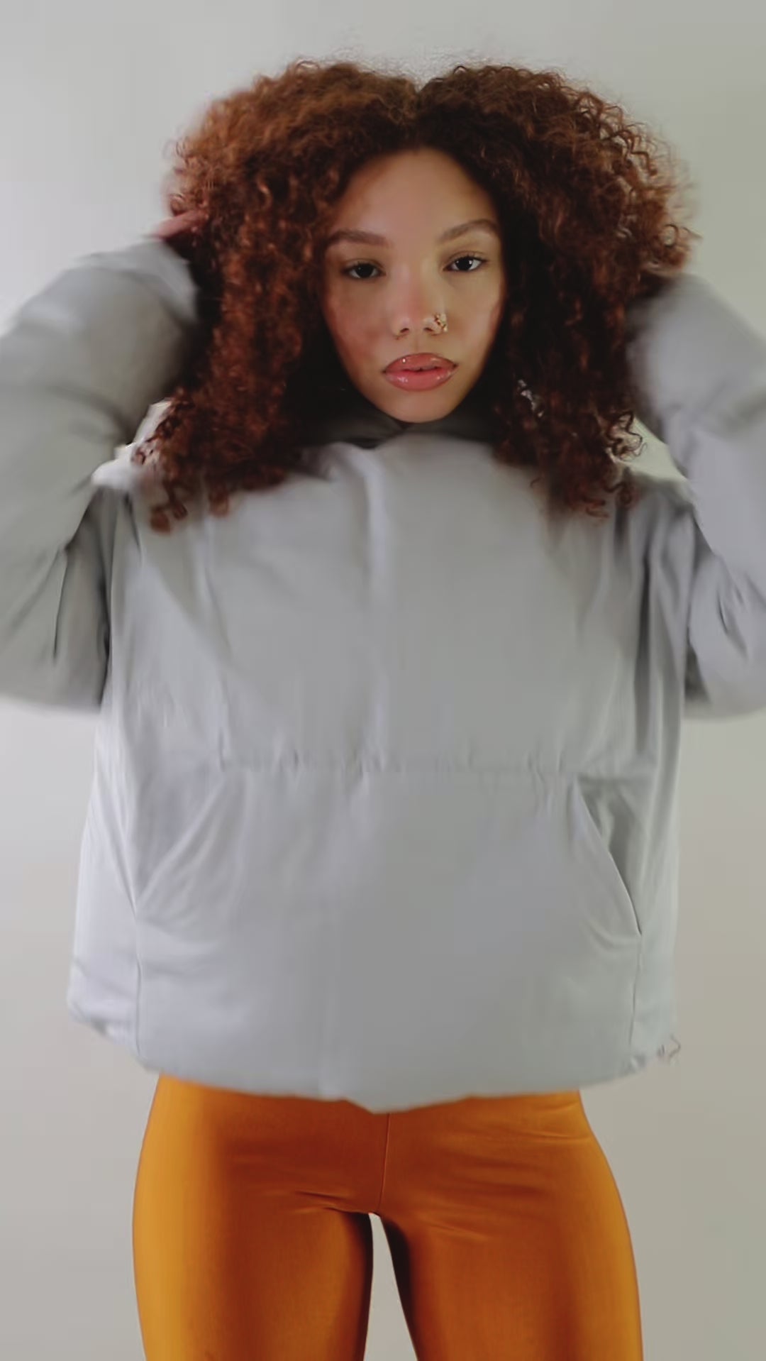 A woman with curly red hair wears a light gray oversized puffer jacket hoodie with a relaxed fit and kangaroo pockets. She pairs it with burnt-orange leggings, standing against a plain white background. The hoodie has a minimal design with a soft, padded texture, ideal for warmth and style