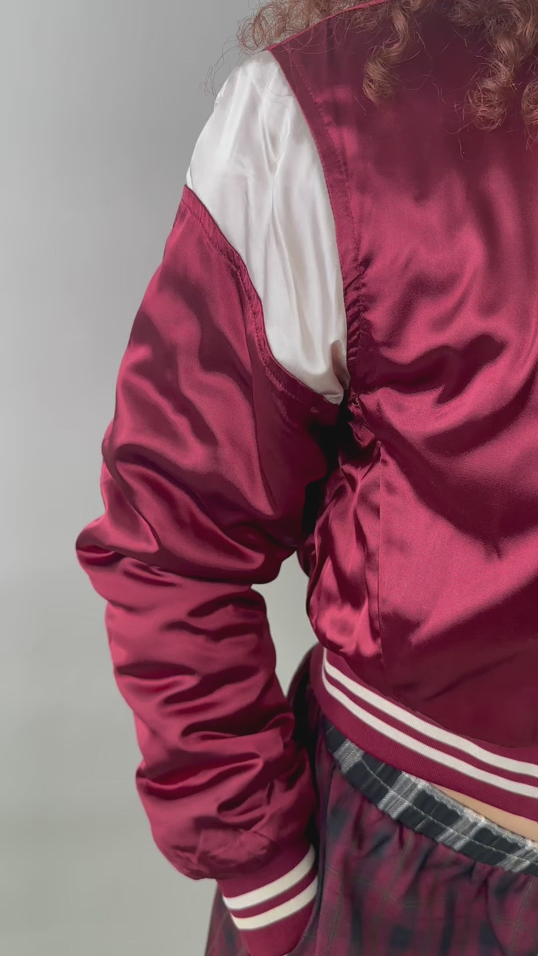 A woman wearing a cropped burgundy satin bomber jacket with white sleeve accents and striped ribbed cuffs. The jacket has a varsity-inspired design with a sleek, shiny finish, giving it a sporty yet stylish look.