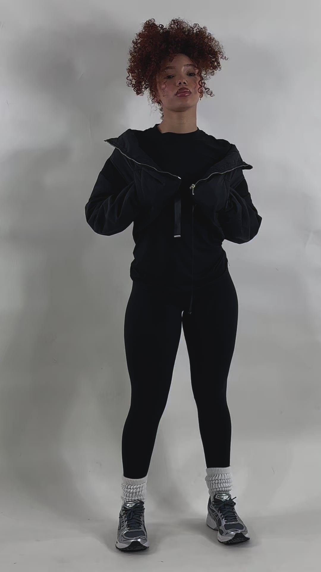 video Woman with curly red hair wearing a brown cropped puffer jacket over a matching brown fitted outfit, posing with hands on the jacket collar against a plain white background. Trendy and cozy outerwear loo