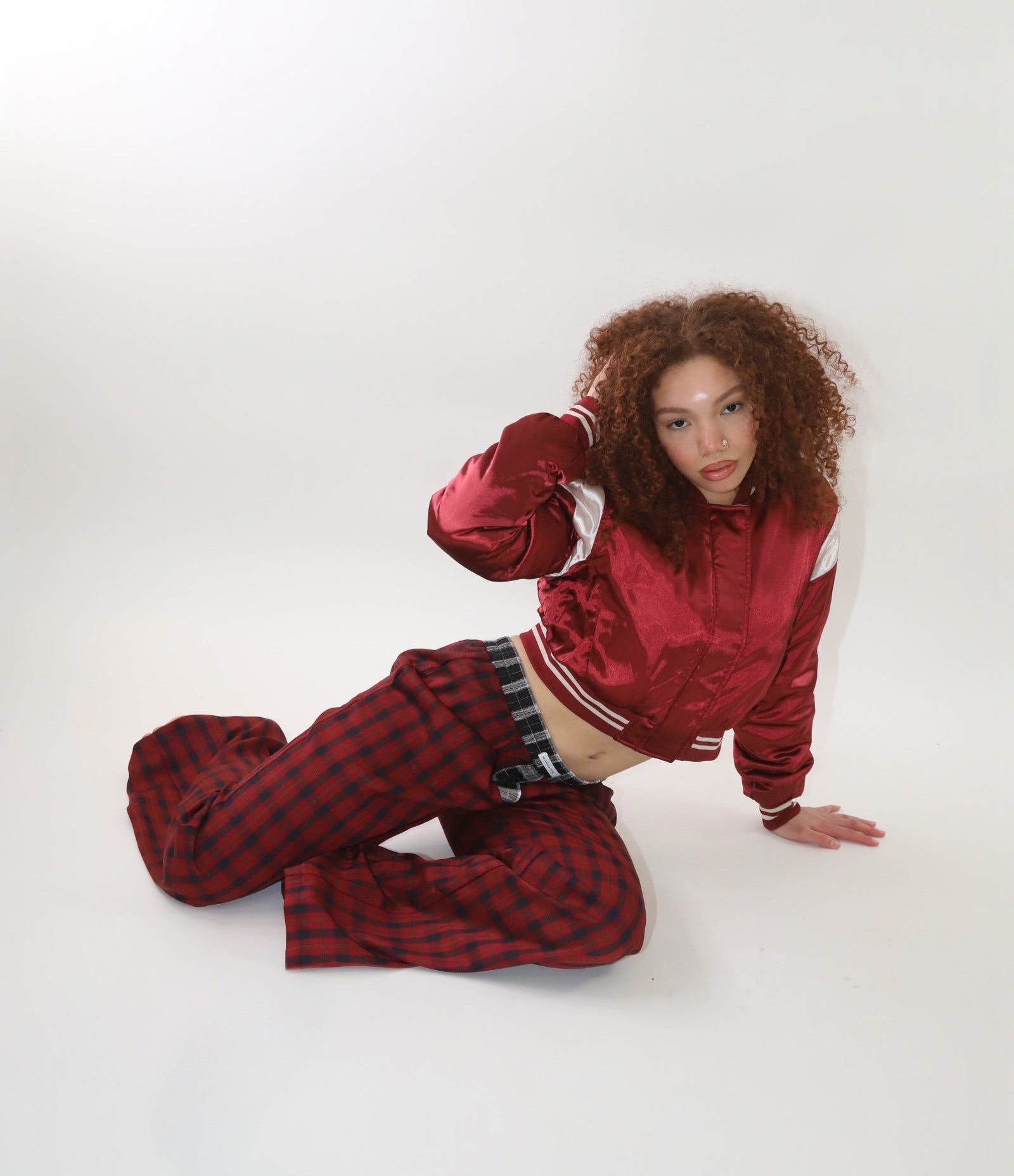 A woman wearing a cropped burgundy satin bomber jacket with white sleeve accents and striped ribbed cuffs. The jacket has a varsity-inspired design with a sleek, shiny finish, giving it a sporty yet stylish look.
