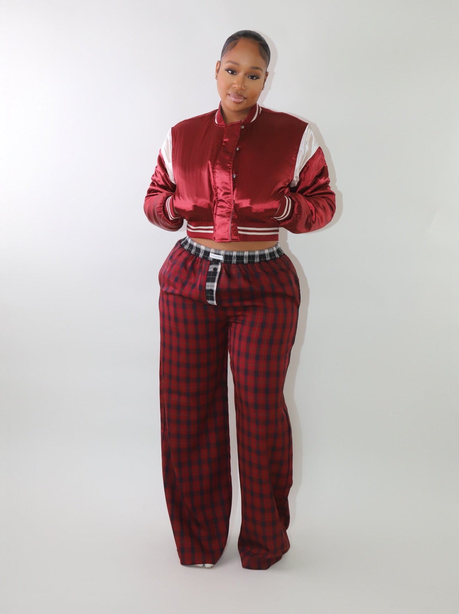 A woman wearing a cropped burgundy satin bomber jacket with white sleeve accents and striped ribbed cuffs. The jacket has a varsity-inspired design with a sleek, shiny finish, giving it a sporty yet stylish look.
